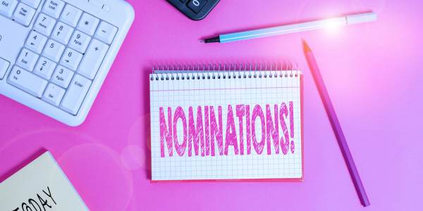 Notebook on a pink desk reading “NOMINATIONS!” with pens and a calculator nearby