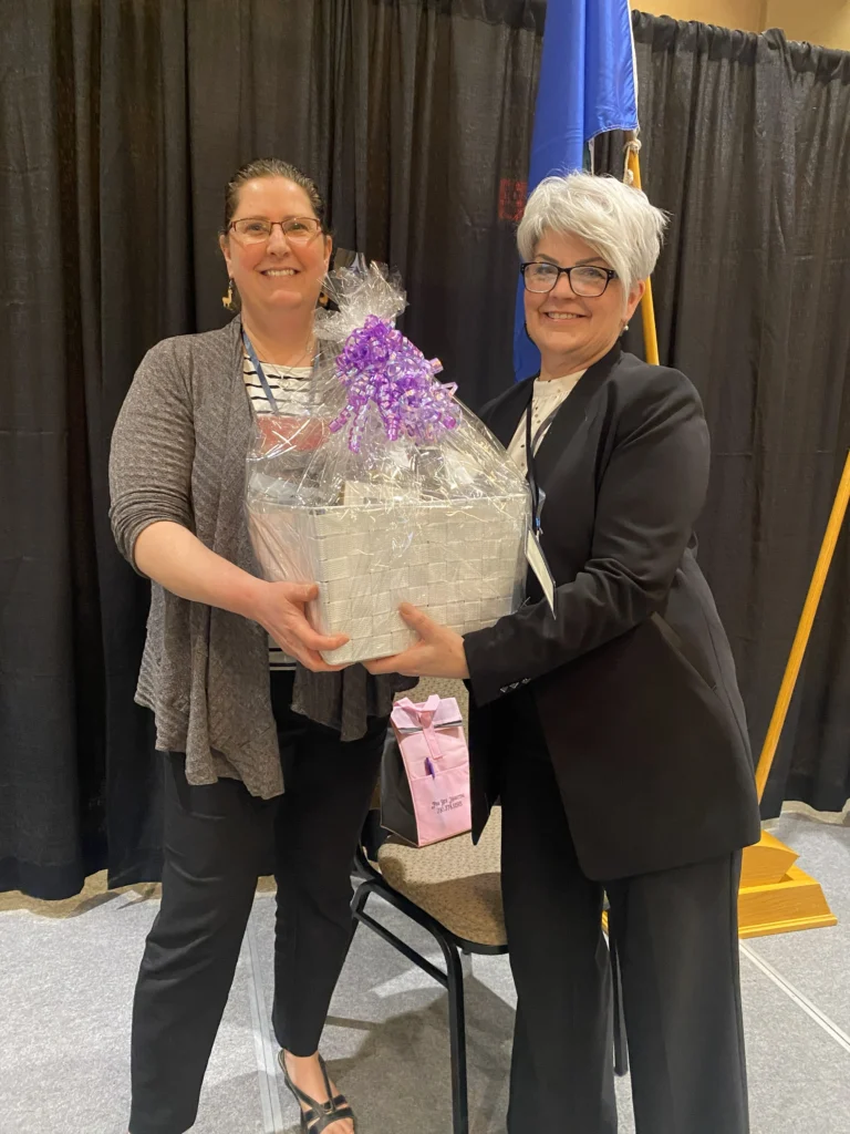 Two women smiling while holding a gift basket in front of a “Shine Bright – Lead Boldly” photo backdrop at the MVAN Women in Business Awards